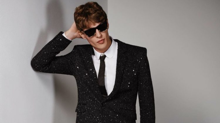 Tim Schuhmacher wears a twill bouclé sequin jacket and pants with a shirt, tie, sunglasses, bracelet, and patent leather oxfords by Saint Laurent.